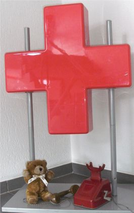 Ein Bärchen sitzt unter einem roten Deko-Kreuz zusammen mit einem alten roten Telefon mit Kurbel